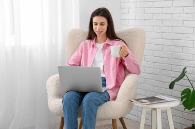 Evdeki ışık perdesinin yanındaki koltukta dizüstü bilgisayar kullanan genç bir kadın.