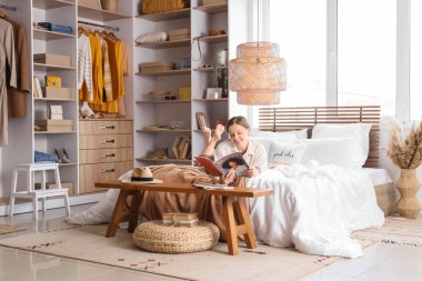 Genç kadın yatakta dergi okuyor. Yatak odasında şık bir gardırobu var.