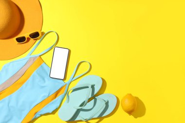 Sarı arka planda modern cep telefonu, mayo ve plaj aksesuarları olan kompozisyon