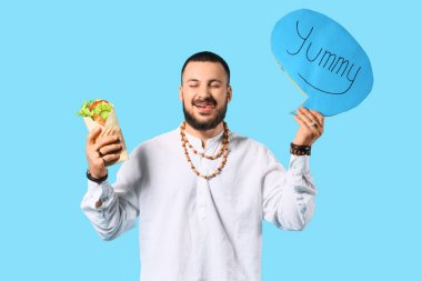 Genç Müslüman bir adam mavi arka planda YUMMY ve sandviç paketiyle konuşma balonu tutuyor.