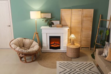 Katlanan perdeli oturma odası, parlayan lambalar, koltuk, hasır masa, kanepe ve mavi duvarın yanındaki şömine.