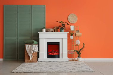 Katlanmış perdeli şömine, oturma odasındaki turuncu duvarın yanında raf ve sepet.