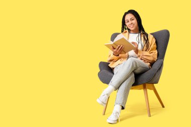 Güzel, mutlu, Afro-Amerikalı bir kadın sarı arka planda koltukta kitap okuyor.