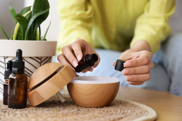 Woman pouring essential oil into air humidifier on table at home, closeup