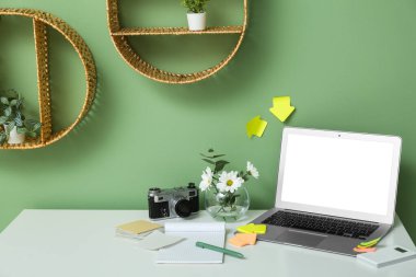 Yeşil duvarın yanında dizüstü bilgisayarı, çiçekleri, kırtasiyesi ve fotoğraf makinesi olan modern ofis işyeri