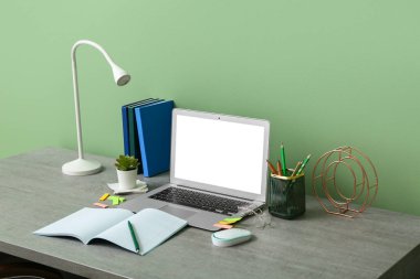 Yeşil duvarın yanında dizüstü bilgisayarı, kırtasiyesi ve lambası olan modern ofis çalışma alanı