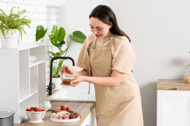 Genç ve güzel bir kadın taze çileği kaseye bandırıyor. Mutfakta erimiş çikolata var.