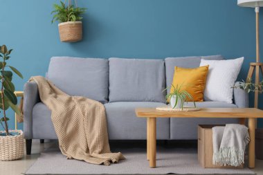 Mavi duvarın yanındaki yastıklı, ekoseli ve kahve masalı rahat bir kanepe.