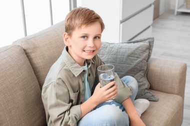 Oturma odasındaki kanepede bir bardak suyla oturan sevimli küçük çocuk.