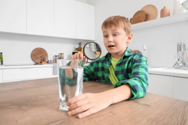Şaşkın küçük çocuk mutfakta büyüteçle su bardağına bakıyor.