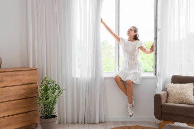 Oturma odasındaki perdelerle pencere eşiğinde oturan genç bir kadın.