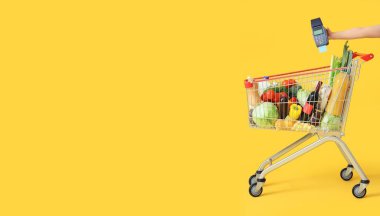 Farklı taze ürünlerle dolu alışveriş arabası ve sarı arka planda el ele tutuşma terminali. Mesaj için yer var.