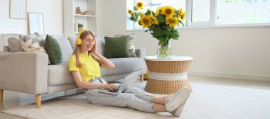 Kulaklıklı, dizüstü bilgisayarlı ve buket ayçiçekli güzel genç bir kadın evde oturuyor.