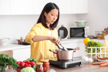 Genç Asyalı kadın mutfaktaki portatif elektrikli ocakta tencerede lezzetli makarna pişiriyor.