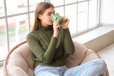 Genç bayan evdeki koltukta sıcak çay içiyor.