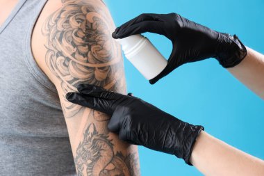 Dövme ustası adamın koluna krem sürüyor, mavi arka planda, yakın plan.