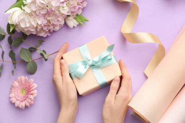 Hediye kutusu olan kadın elleri, ambalaj kağıdı ve mor arka planda güzel çiçekler. Uluslararası Kadınlar Günü