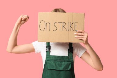 Protesto eden kadın inşaat işçisi üzerinde yazı olan bir pankart tutuyor ve pembe arka planda kasları gösteriyor.