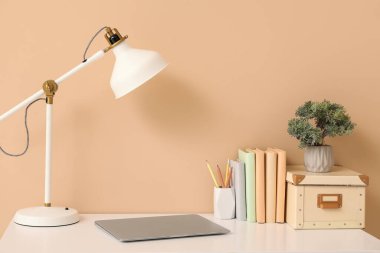 Bej duvarın yanında lamba, kitap ve dizüstü bilgisayarı olan şık bir iş yeri.