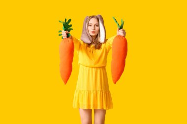 Sarı arka planda oyuncak havuçları olan tavşan kulaklı güzel bir kadın. Paskalya kutlaması