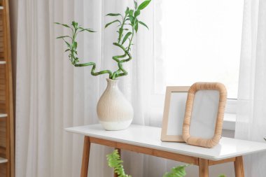 Yatak odasının penceresinin yanındaki masada bambu saplı vazo ve fotoğraf çerçeveli.