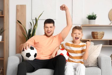 Genç adam ve küçük oğlu evde futbol toplarıyla futbol takımı için tezahürat yapıyorlar.