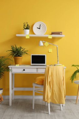 Çalışma odası, dizüstü bilgisayar, sandalye ve saat sarı duvarda.