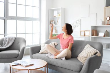 Oturma odasındaki kanepede oturan klimalı uzaktan kumandalı gülümseyen genç kadın.