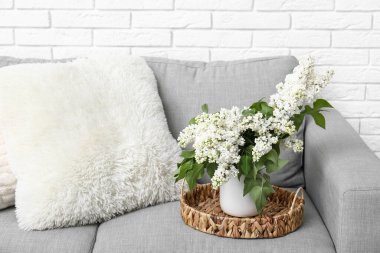 Oturma odasındaki beyaz tuğla duvarın yanındaki kanepede çiçek açan leylak dallı vazo.