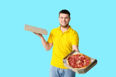 Elinde karton kutular ve mavi arka planda lezzetli pepperonili pizzalar tutan genç adam.