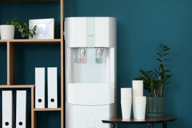 Masasında kağıt bardaklar ve mavi duvarın yanında raf ünitesi olan modern su soğutucusu.