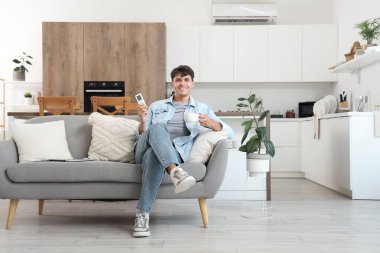 Klimalı, uzaktan kumandalı ve mutfaktaki kanepede oturan genç bir adam.