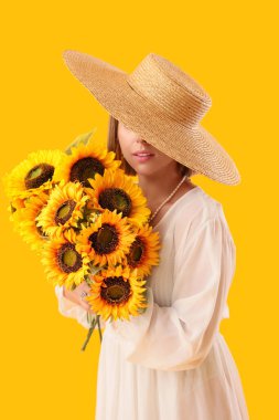 Hasır şapkalı, şık makyajlı ve sarı arka planda bir buket ayçiçeği olan güzel bir kadın.