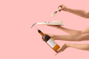 Pembe arka planda tirbuşonlu ve peynirli şarap şişesini tutan kadın eller.