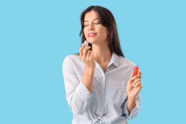 Güzel genç bir kadın mavi arka planda dudaklarına sıvı ruj sürüyor.