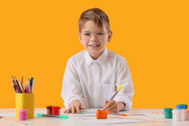 Küçük çocuk sarı arka planda malzemelerle masada çizim yapıyor.