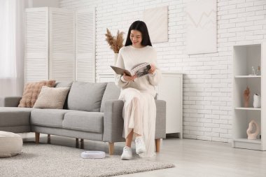 Evde elektrikli süpürgeyle kanepede dergi okuyan genç bir kadın.