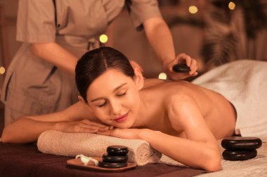Koyu renk spa salonunda genç bir kadın sıcak taşlarla masaj yaptırıyor.