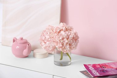 Ortanca çiçekli vazo, dergiler ve pembe duvarın yanındaki çekmecelerde çaydanlık.
