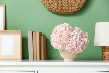 Yeşil duvarın yanındaki rafta pembe ortanca çiçekleri ve kitapları olan vazo.