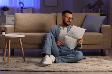 Gözlüklü yakışıklı adam, akşam evde halıda gazete okuyor.