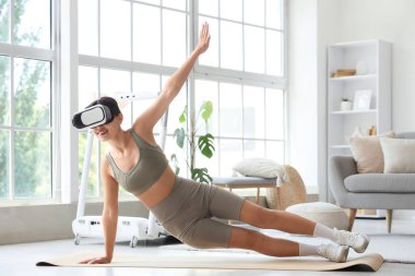 VR gözlüklü genç bir kadın evde yoga minderiyle egzersiz yapıyor.