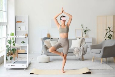 Sporty young woman in VR glasses practising yoga at home