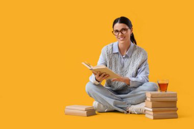 Kitapları ve fincanı olan güzel bir kadın sarı arka planda oturuyor.