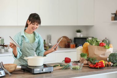 Tablet bilgisayarlı genç bir kadın mutfakta çorba yapıyor.