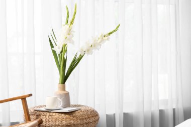 İçinde gladyatör çiçekleri olan vazo, bardak ve kitap odadaki ışık perdesinin yanındaki masada.