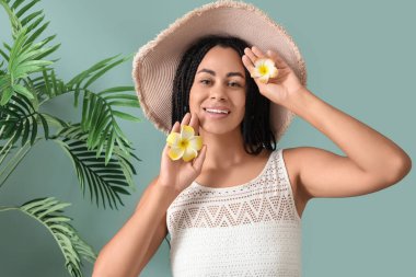 Plumeria çiçekleri ve yeşil arka planda palmiye yaprakları olan şapkalı güzel bir Afro-Amerikalı kadın.