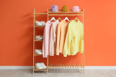 Turuncu duvarın yanında kapüşonlu, spor ayakkabılı ve şapkalı raflar.