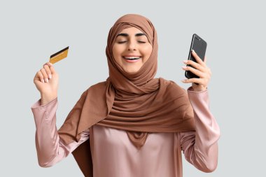 Gri arka planda cep telefonu ve kredi kartı olan mutlu Müslüman kadın.