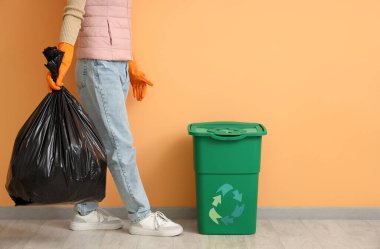 Turuncu duvarın yanında çöp torbası ve çöp kutusu olan bir kadın.